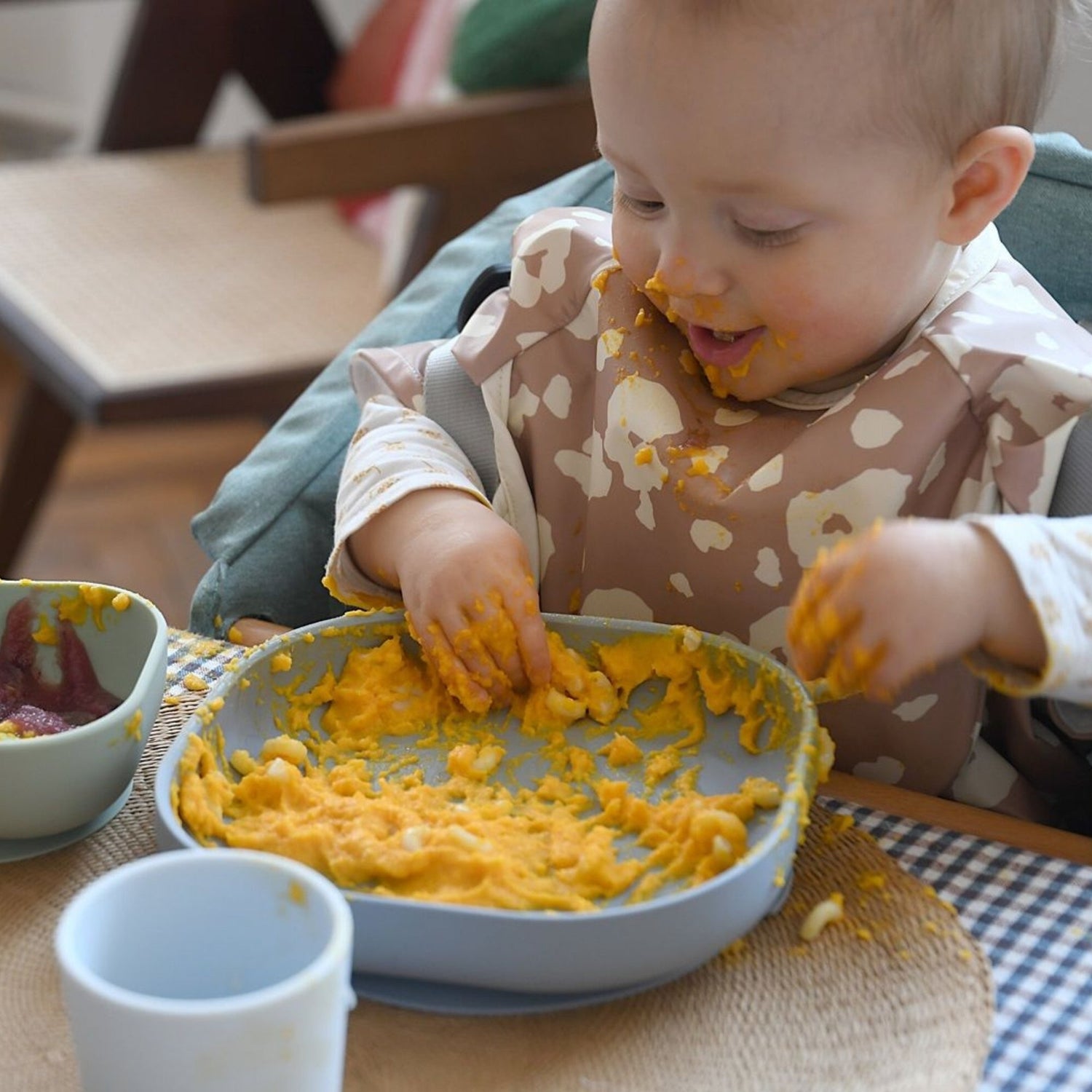 baby sitting in high chair eating orange puree from a grey silicone bowl with his hands